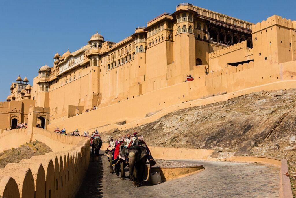 A majestic view of the Amber Fort in Jaipur, Rajasthan, India. Ornate elephants adorned with colorful fabrics carefully descend the fort's ramp, carrying tourists on a unique journey. Experience the grandeur of Amber Fort with our ONE DAY DELHI TO JAIPUR TRIP PACKAGE.
